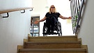 Kerstin Rathgeb im Rollstuhl. Unten wäre die Toilette: Eine Treppe aber versperrt für Kerstin Rathgeb, die im Rollstuhl sitzt, den Weg. | Bild: BR / Kontrovers | Judith Zacher